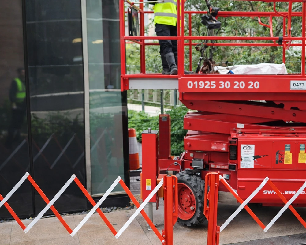 Operative working from a red scissor lift carrying out façade maintenance next to a glazed building exterior with safety barriers in place