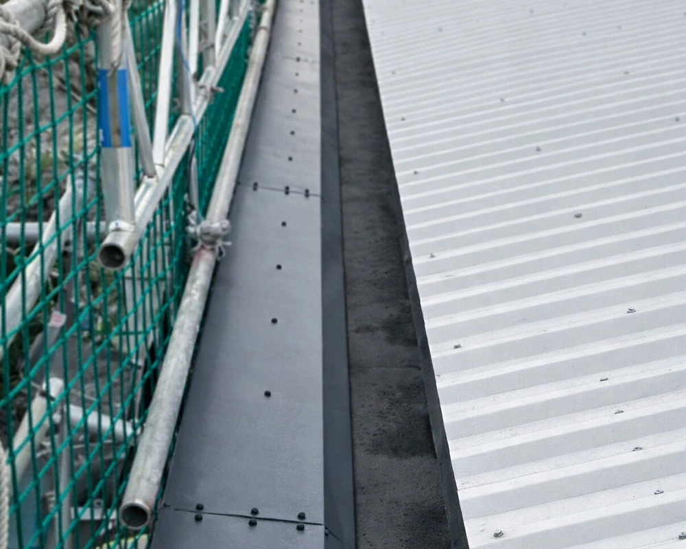 Roof edge detail showing metal flashing, corrugated roof sheets, and temporary scaffold safety netting during maintenance works